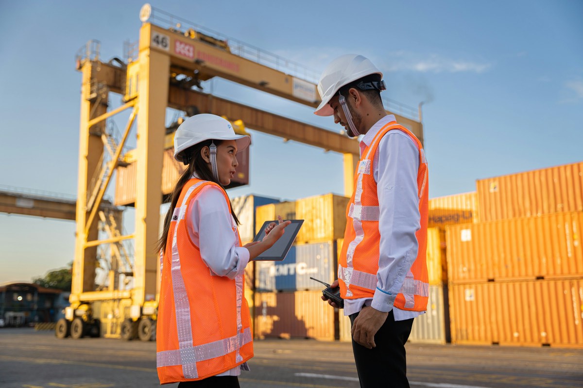Two workers in hard hats discussing plans beside shipping containers
